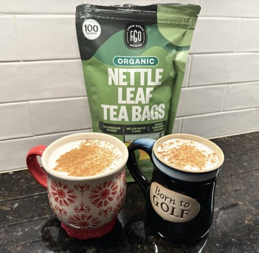 Two mugs of frothed nettle tea with cinnamon and a nettle tea bag on a kitchen counter