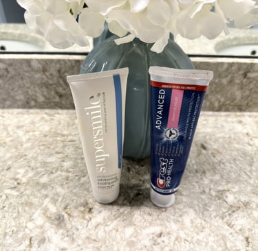 Two tubes of toothpaste on a clean bathroom counter beside a vase of flowers in soft natural light