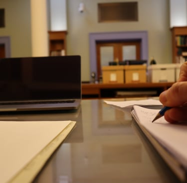 View of a desk in an archival research room showing a person writing in a notebook.