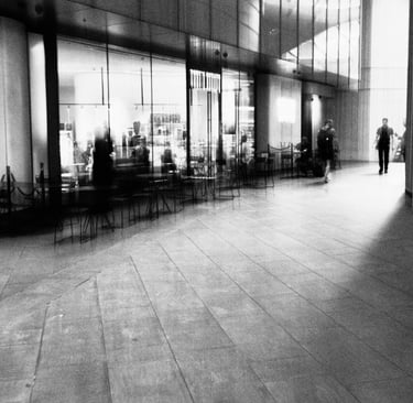 A modern cafe with people moving inside. A lone figure walks on a wide, empty tiled hall, lending a serene urban atmosphere.