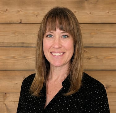 Professional headshot of a smiling woman with brown hair and bangs against a rustic wood wall background.