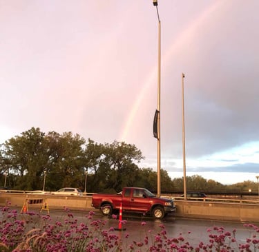 Red truck on a bridge with a beautiful rainbow in the sky, there are trees and flowers