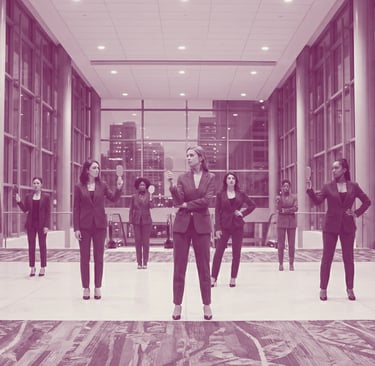 A diverse group of women in colored business suits strike a pose, holding up hand mirrors