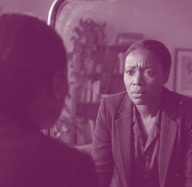 A Black woman in a blue blazer looks intensely at her reflection in an antique mirror