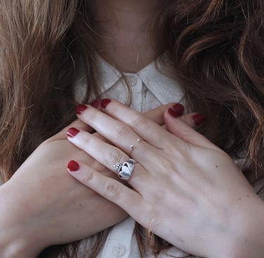A white woman with brown hair clasping her hands over her heart. She's wearing a white blouse and her nails are painted red.