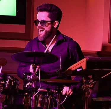 Singer Leo Couto playing the drums, wearing a black shirt, a vest, and sunglasses.