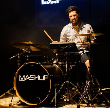 Drummer Lucas Dias wearing a short-sleeved white shirt and a pork pie hat, playing a black drum set.
