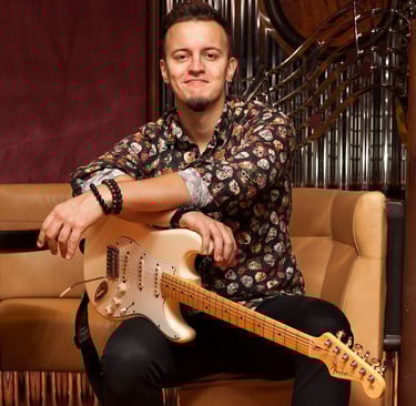 Guitarist Lucas Vianna sitting on a couch with his white Fender Stratocaster guitar on his lap, wearing a colorful shirt and
