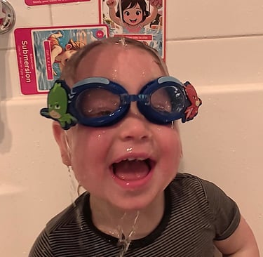 Baby sitting in the bath with goggles with water over the face and swim skill flash cards stuck on the wall.