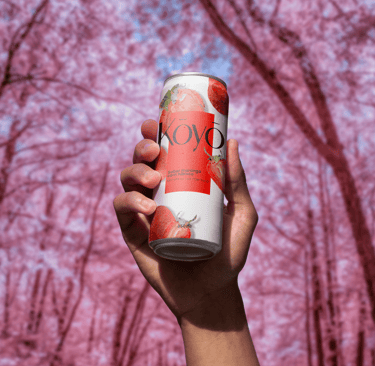Lata da bebida Koyō sabor morango sendo segurada por uma mão, com fundo rosa de árvores floridas, tr