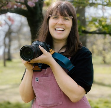 Laura Franklin holds her camera up as she smiles to the camera. It is a London Blossoms photo shoot 
