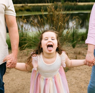 Family photo of a little girl holding her parents hands, she sticks her tongue out at the camera
