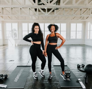 Two smiling women in black activewear standing on yoga mats in a bright fitness studio with gym equipment.