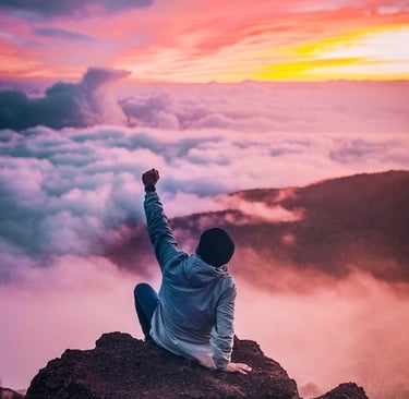 Young man on top of a mountain, above the clouds, with the sun setting in the background.