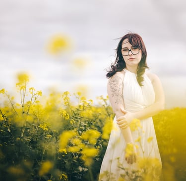 A woman with glasses and a white dress posing in a blooming yellow wildflower field.