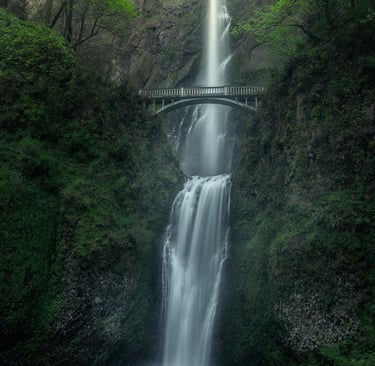 a waterfall cascading down a green gorge and under a walking bridge