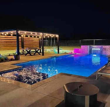 Modern luxury backyard pool with blue LED lighting, a waterfall feature, and a lit pergola at night.