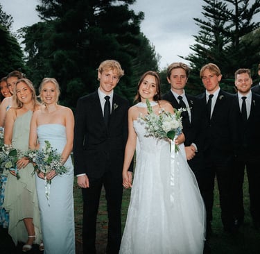 A bride and groom posing outdoors with their wedding party in light blue dresses and black suits.