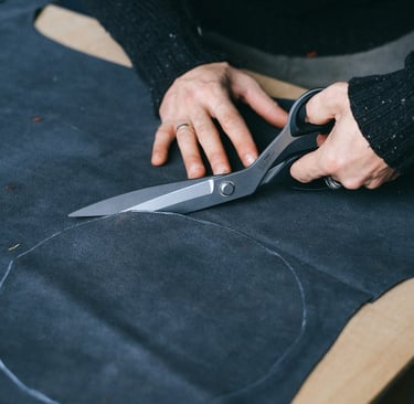 Hands cutting a circular fabric patch from dark denim using large fabric scissors.