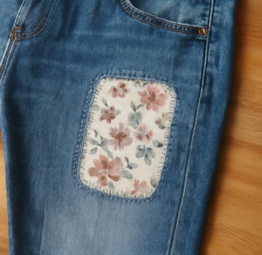 Blue jeans with a rectangular floral patch stitched on the left leg,.