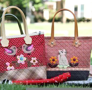 Two quilted fabric tote bags with appliques displayed on grass during a picnic setup.