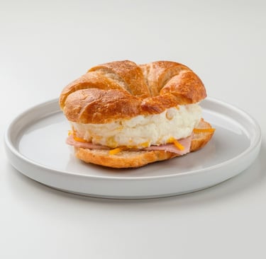 Breakfast croissant sandwich with fluffy eggs, melted cheese, and ham on a white plate.