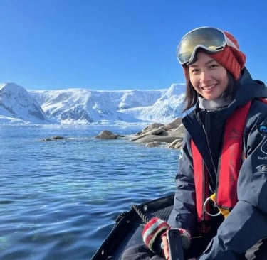 A smiling woman in polar gear on a boat in Antarctica with icy mountains in the background.