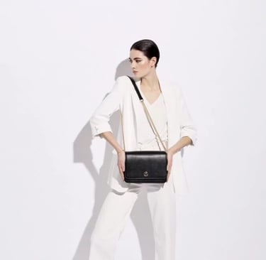 A woman in a white suit holding a black leather crossbody bag with a gold chain strap.