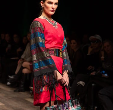 A model walks a fashion runway wearing a vibrant red dress with embroidered fringe sleeves and a woven tote bag.