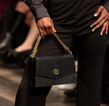 Model holding a black leather designer handbag with a gold chain strap at a fashion show runway.