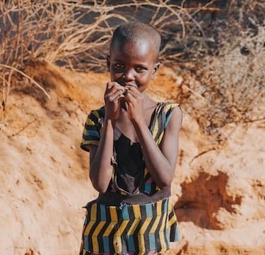A young African girl in a patterned dress stands in a sunny, dry landscape.