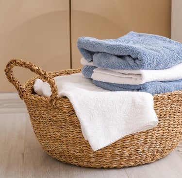 Stack of folded plush blue and white bath towels inside a woven wicker basket on a clean, light hardwood floor. 