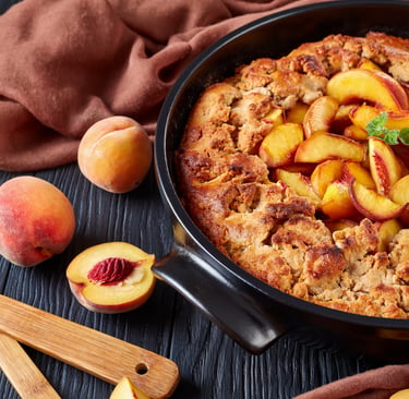 Freshly baked warm peach cobbler in a black skillet with sliced fresh peaches on a dark wooden background.