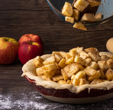 Diced spiced apples being poured from a bowl into an unbaked pie crust next to whole red apples on dark wood. 