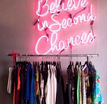 Letrero de neón rosa que dice “Cree en segundas oportunidades” sobre rack de ropa de segunda mano.