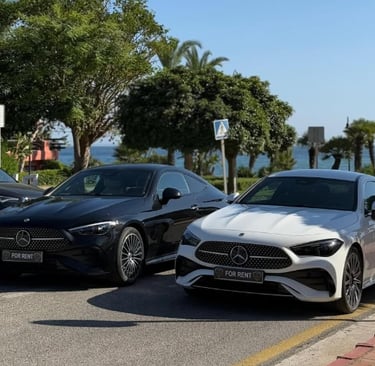 Black and white Mercedes-Benz luxury rental cars parked on a sunny coastal road.