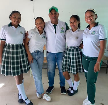 Smiling students in green plaid school uniforms pose with a male mentor in a white classroom.