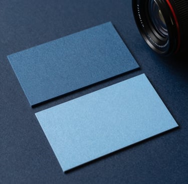 A close-up photograph of high-quality, textured business card samples in Muted Slate Blue and Soft Sky Blue on a clean Dark Navy surface. Professional studio lighting in a modern International / Western design office, showcasing a clean and sophisticated aesthetic.