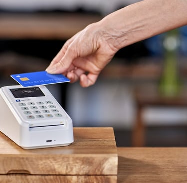 Customer making a contactless credit card payment on a white SumUp card reader at a retail counter.