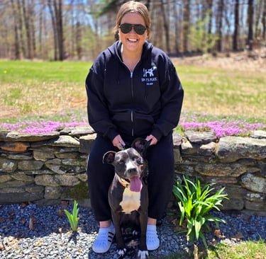 A smiling woman in sunglasses sits on a stone wall with a happy black and white dog. 