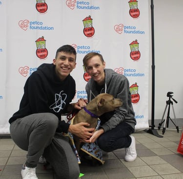 Two smiling individuals crouch beside a brown dog in front of a Petco Foundation backdrop.
