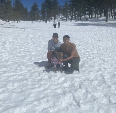 Two men crouched in a snowy field, smiling at the camera with a dog in a pink jacket between them.