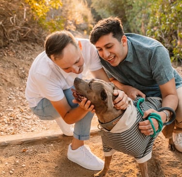 Two people crouch on a dirt path, affectionately petting a dog in a striped sweater.