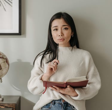woman holding a pencil and red notebook, studying thoughtfully in a minimalist workspace.