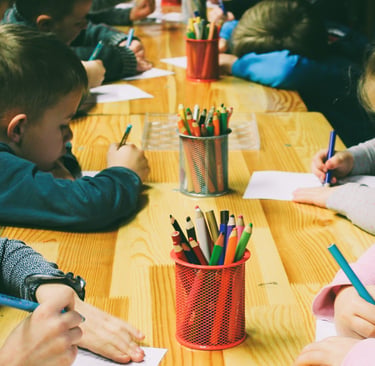 Children are gathered around a wooden table, drawing with colored pencils. 