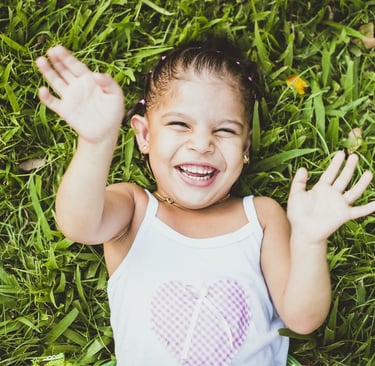 A joyful child in a white heart-patterned shirt lies on grass, smiling broadly with arms raised.
