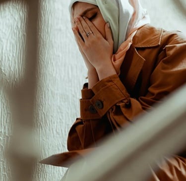 A woman in a hijab and brown coat sits on stairs covering her face in distress or prayer.