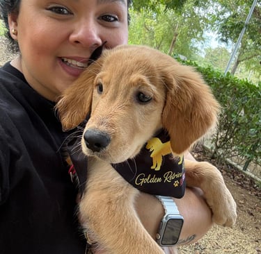 A smiling woman holding a fluffy Golden Retriever puppy wearing a black bandana outdoors.