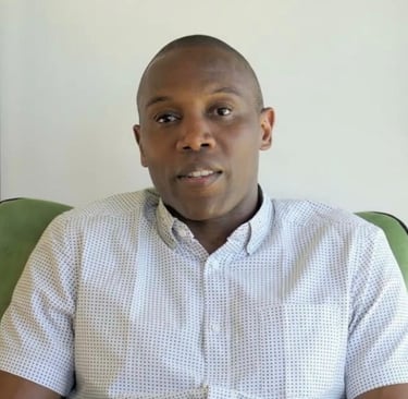 Portrait of a Black man in a patterned short-sleeve button-down shirt sitting on a green chair.