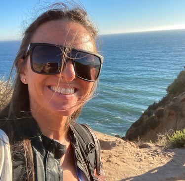 Michael Alexa Pitts smiling in the sunshine wearing black shades, a rocky Pacific coastline, blue sky & blue ocean behind